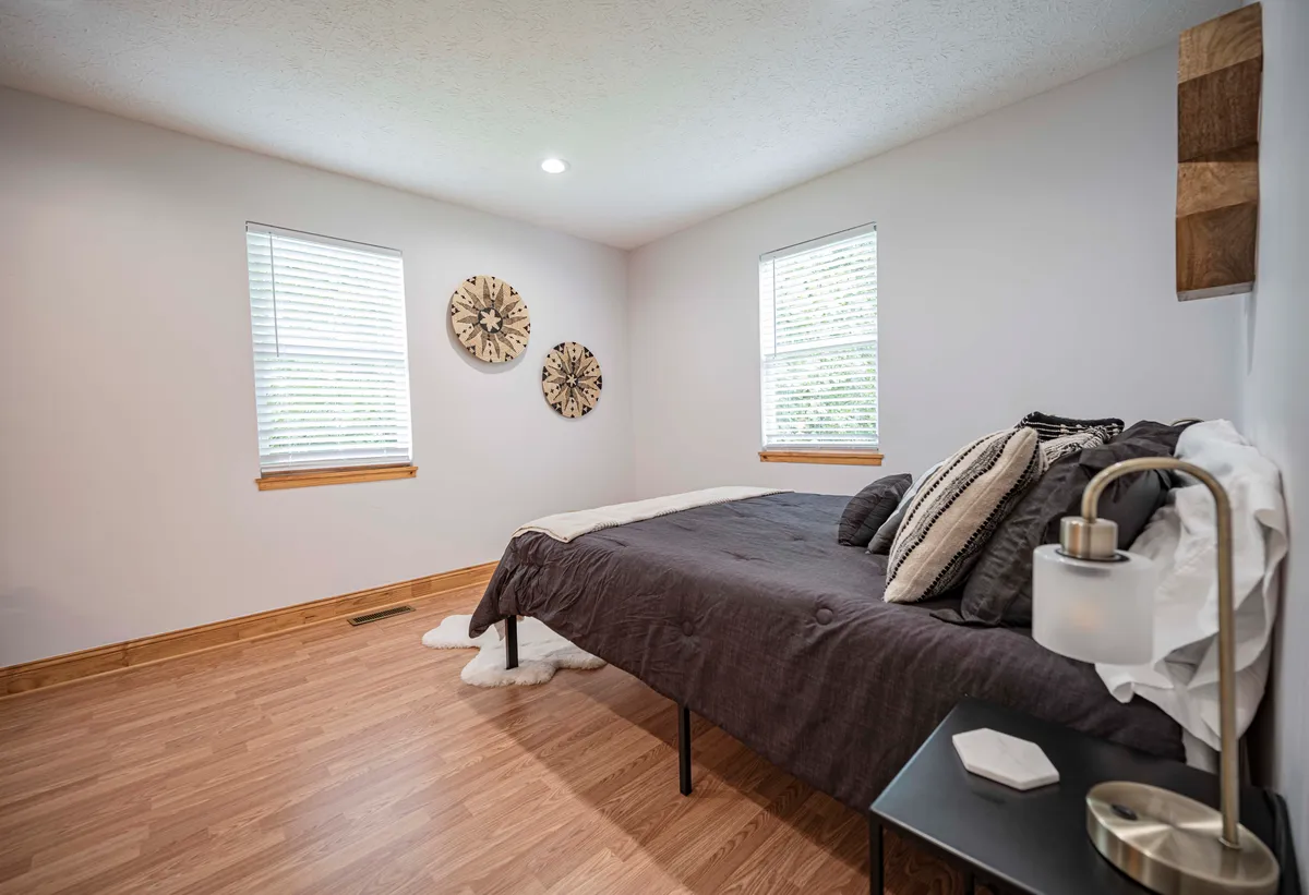 A cozy, serene bedroom interior with a bed dressed in dark bedding, illuminated gently by sunlight. Decorative wall clocks and a nightstand with a lamp add an intimate, inviting touch, ideal for restful mornings or peaceful evenings.