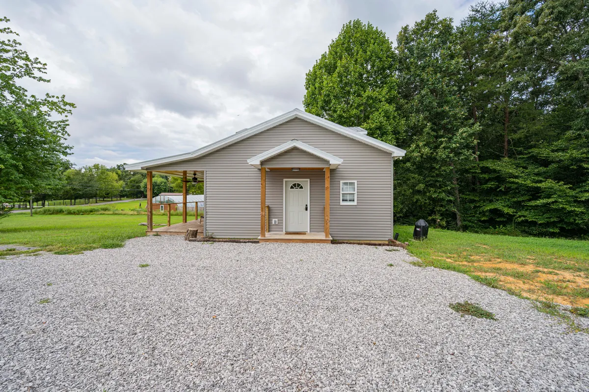 A welcoming view of the front porch with sleek cable railing, framed by green lawns and mature trees. The gravel driveway offers a sense of spaciousness for parking, while the porch itself is ready for gatherings or quiet moments. The home exudes a country charm and embraces the natural surroundings.