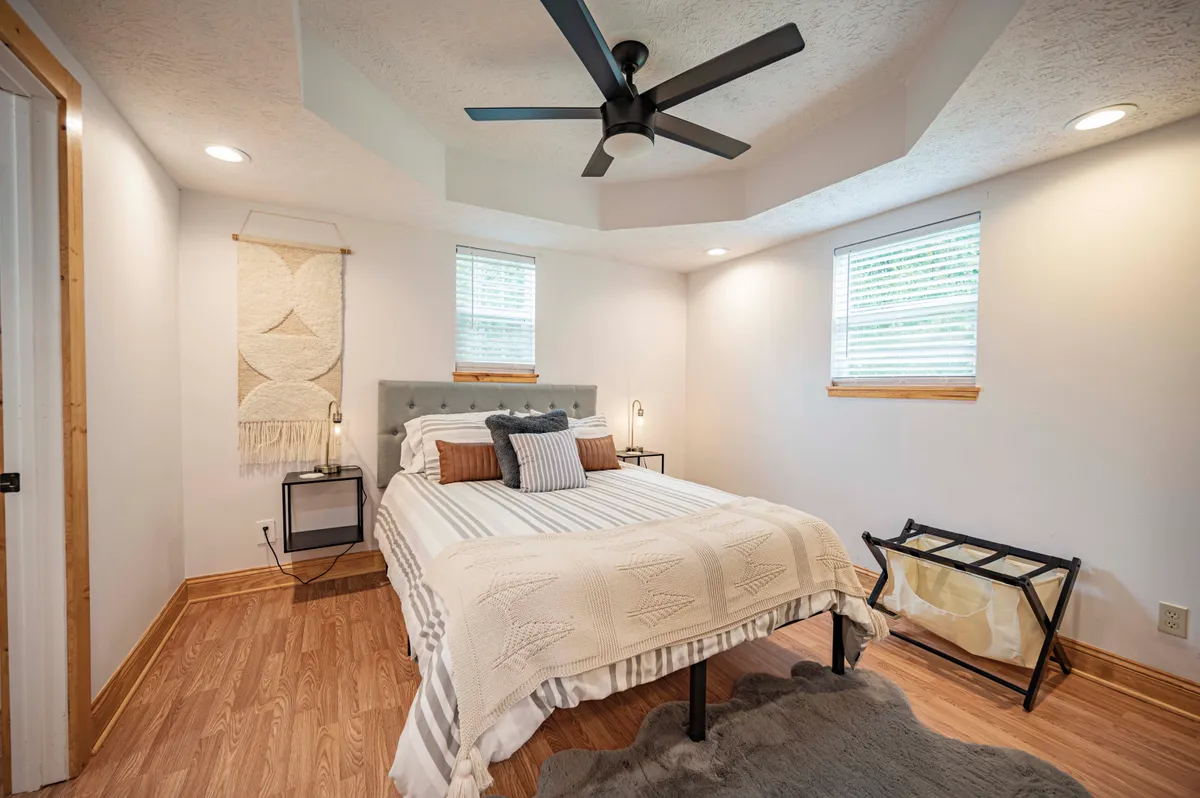 A serene bedroom with a cozy bed topped with plush bedding, a textile wall hanging above the headboard, large windows letting in natural light, and a ceiling fan overhead. The room also features a reading nook by the window, creating a peaceful and inviting atmosphere.