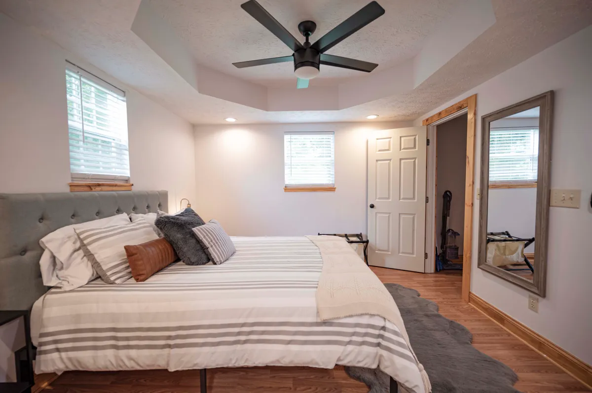 A peaceful bedroom featuring a neatly made bed, large windows letting in ample natural light, a fan for comfort, and a mirror that reflects the serene, uncluttered atmosphere. The space feels inviting and restful, ideal for private retreats after a productive workday.